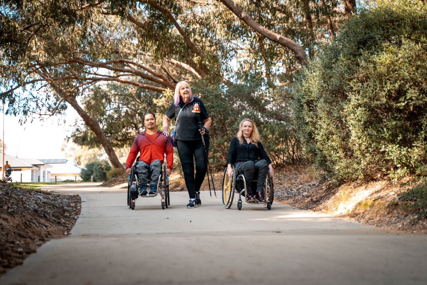 Three people walk and roll together down a wide, paved accessible path through a sun-dappled park surrounded by large native trees and shrubs.